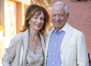 A smiling woman with brown hair wearing a gray shirt and coat stands beside a smiliing man with white hair wearing a blue shirt and white sport coat.
