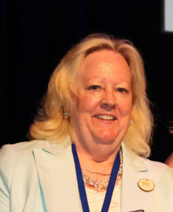 A smiling woman with blonde hair wearing a beige suit coat.