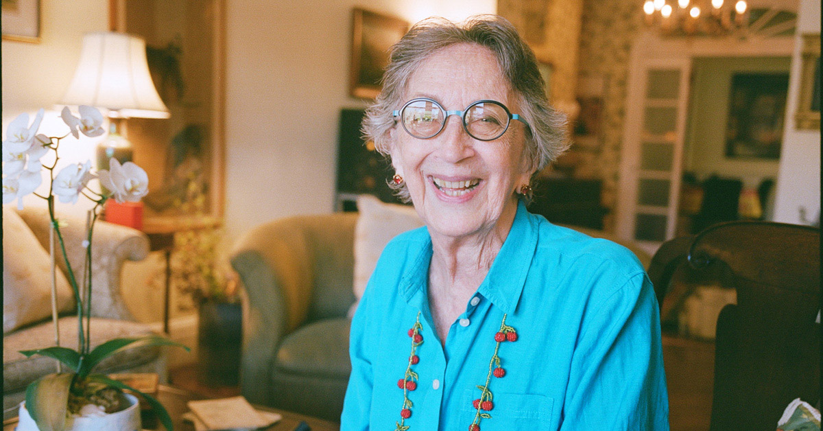 A smiling elderly woman with gray hair wearing glasses and a light blue shirt sits in a living room.