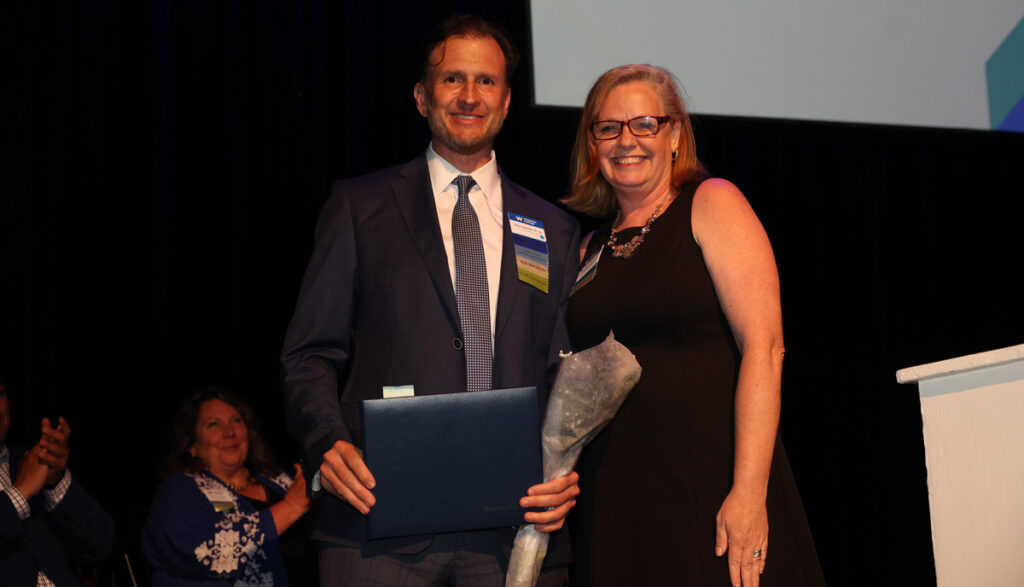 A smiling man wearing a darks suit, white shirt and tie stand next to a smiling woman wearing a black dress.