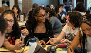 In a room of young men and women seated at tables, a Black woman wearing glasses and a black shirt looks at a white young woman with dark hair wearing a white T-shirt.