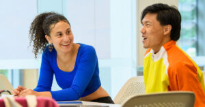 A smiling young woman in a blue shirt looks at a smiling young man wearing an orange and yellow shirt as they sit at a table together.