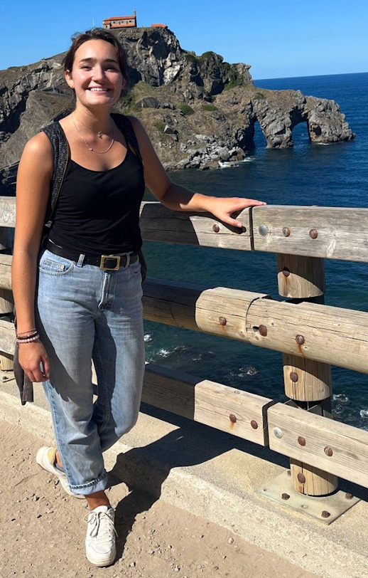 A smiling young woman with dark hair wearing a black tank top, blue jeans and white sneaker rests her left arm on the railing of a fence. The water from a sea or ocean and rock formations are visible in the background.