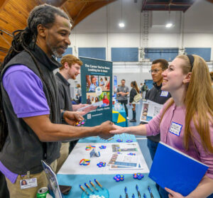 A smiling tall Black man wearing a purple short-sleeved shirt and a dark vest shakes the hand of a young white woman with blonde hair wearing a pink shirt and holding a blue folder in her left arm.