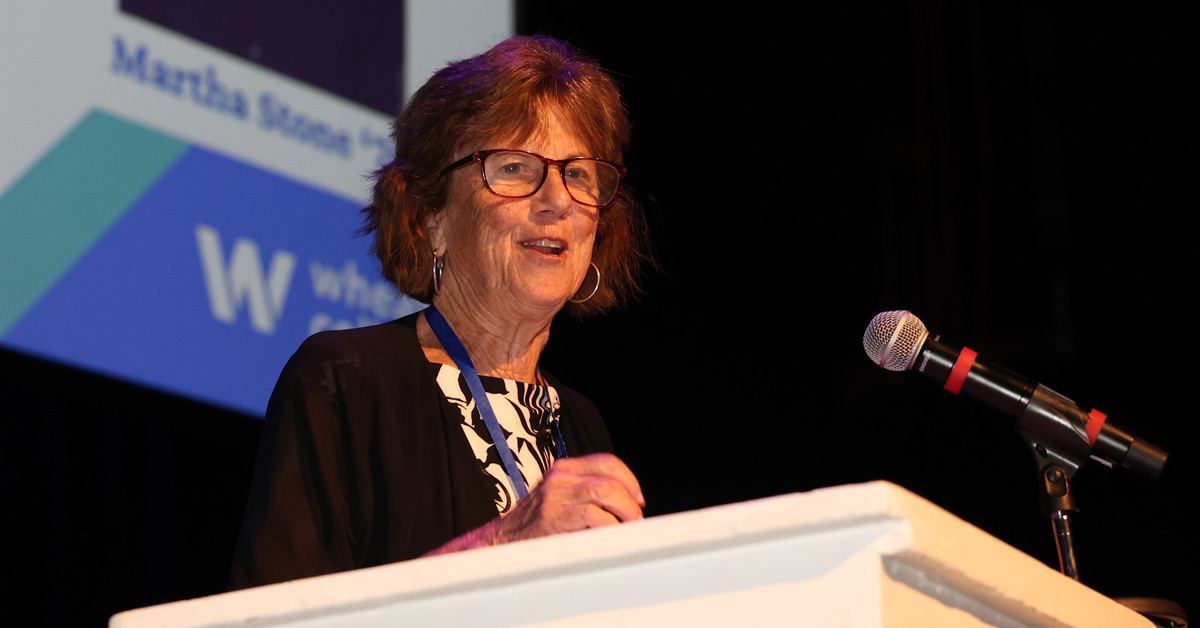 A woman with short brown hair wearing glasses and a dark suit coat speaks from a podium.