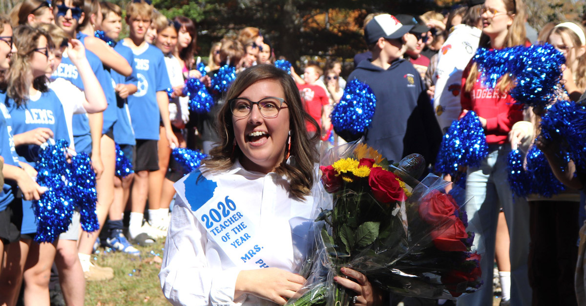 A smiling woman with brown hair wearing glasses, a white shirt and a sash reading "2026 Teacher of the Year" holds bouquets of flowers, walking past middle school students.