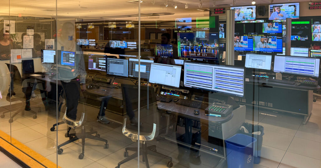 A room of television monitors on desks and mounted from ceilings behind a wall of glass displays various programs and information.