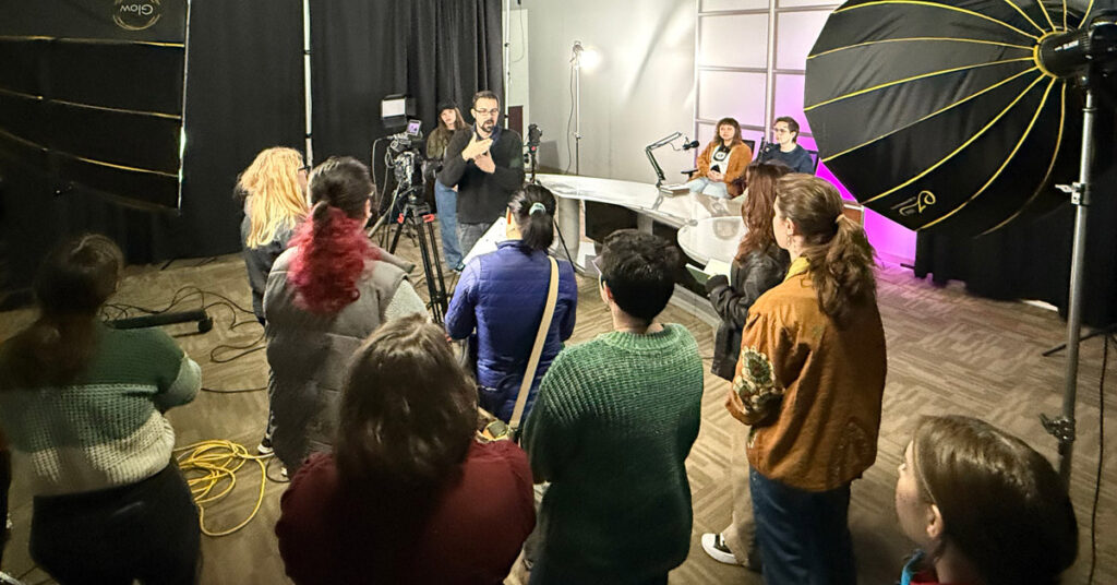 A man addresses a crowd of young men and women in a studio with lighting and camera equipment.