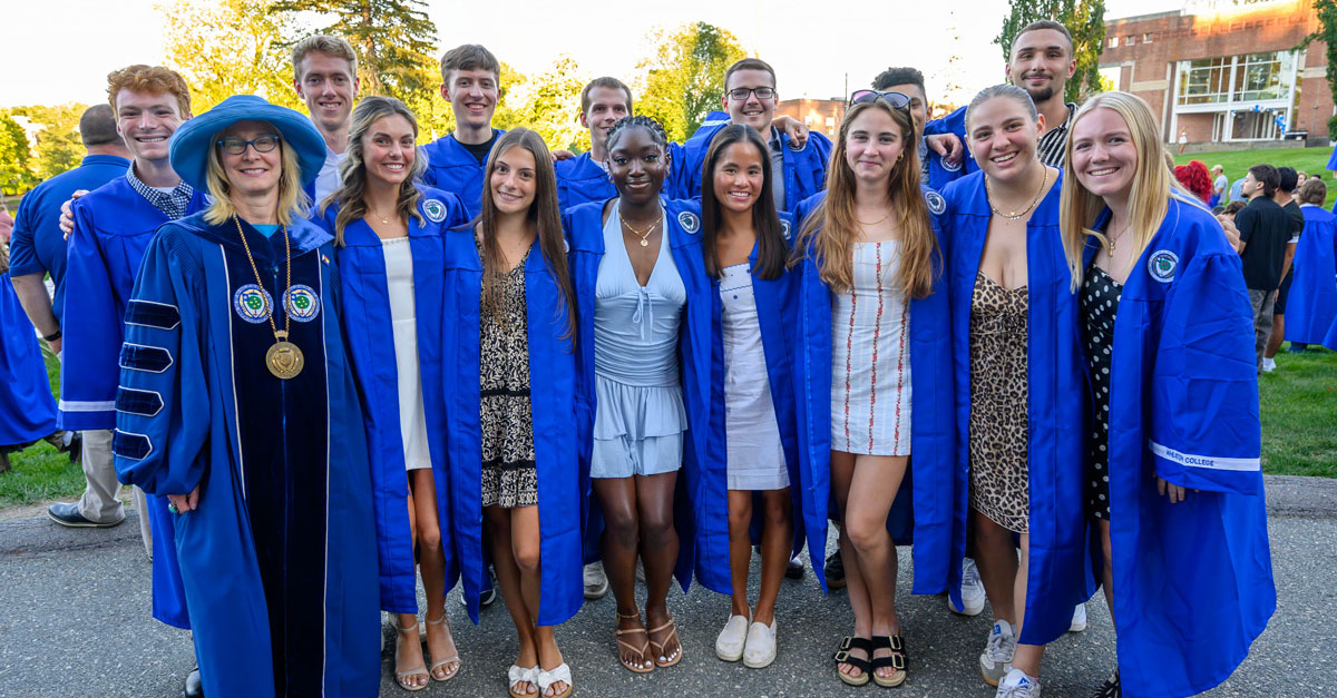A group of 15 individuals, all wearing royal blue academic robes, pose for an outdoor photo.