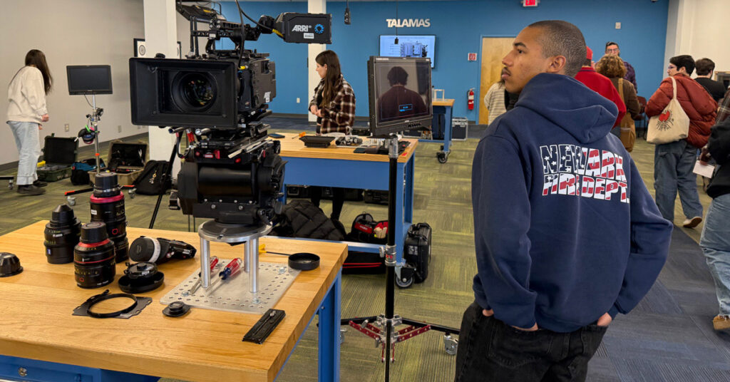 An individual wearing a hooded blue sweatshirt and black pants stands beside a table in a room filled with camera equipment.