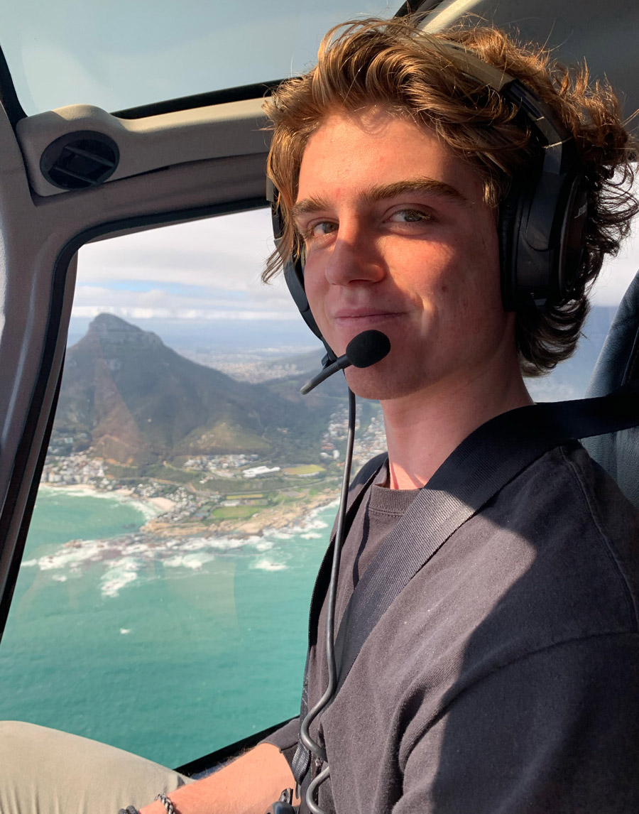 A young man with wavy brown hair wearing a dark T-shirt and a headset with a microphone sits inside a helicopter with the greenish ocean and mountains visible below outside the window behind him.