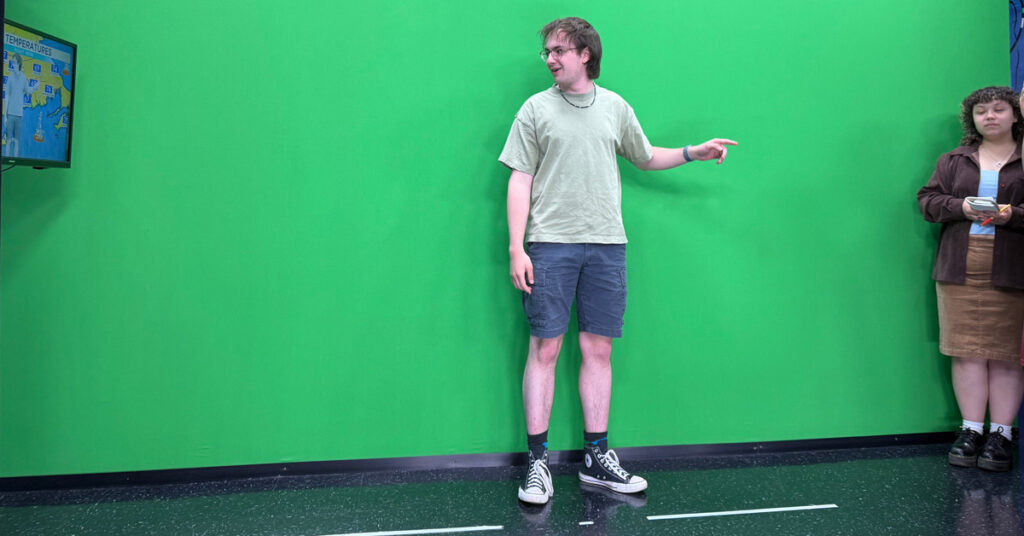 A young man wearing a light T-shirt and dark shorts stands in front of a green-colored wall while looking at a TV monitor to his right.