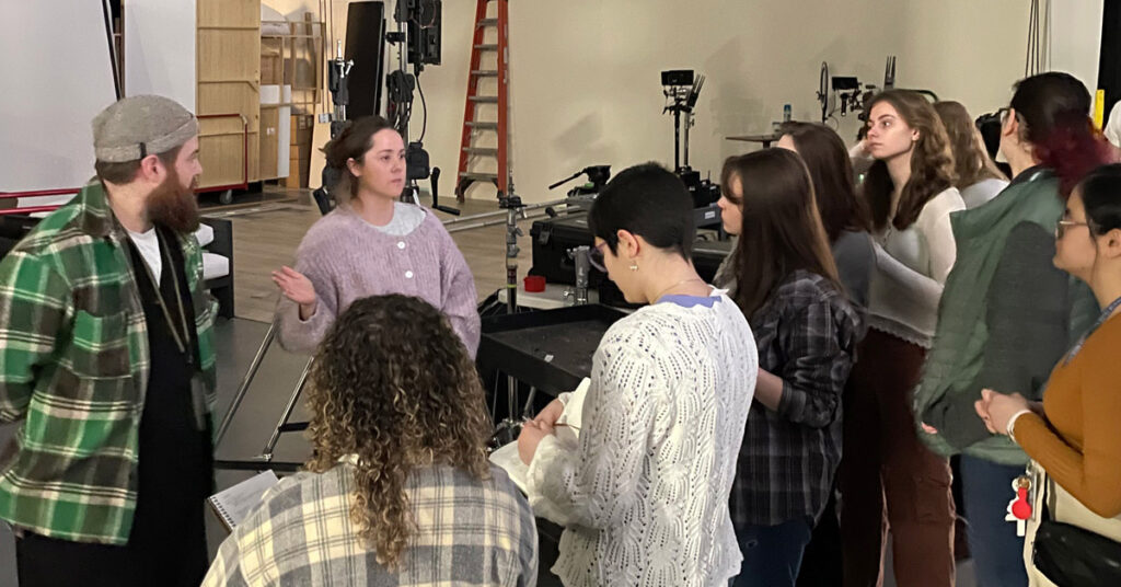 A woman with dark hair and a gray shirt talks to a group of young men and women in a room.
