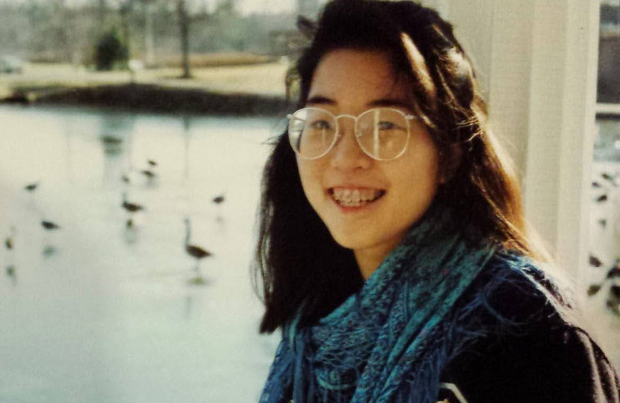 A smiling young woman with dark hair wearing glasses a dark coat and scarf stands outside. A frozen pond with geese standing on it is in the background.