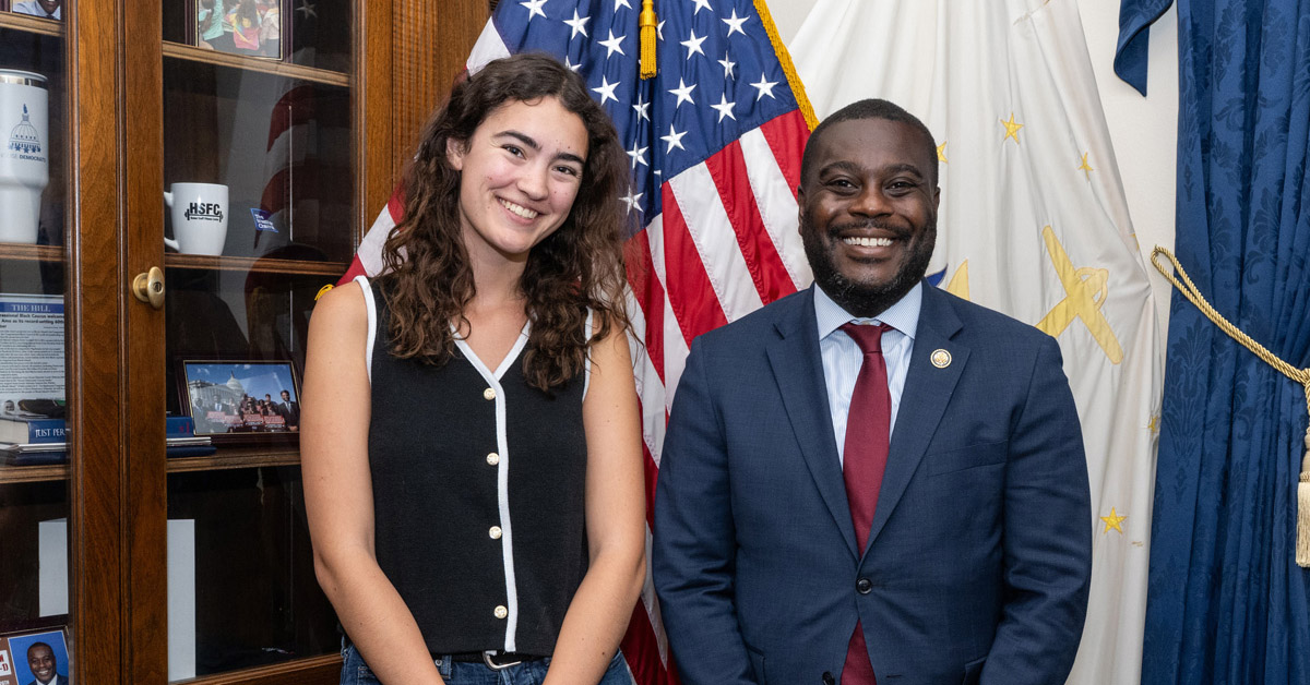 A smiling young woman with long dark wearing a black button-down tank top stands beside a smiling Black man wearing a dark blue suit and a red tie in fron of an American flag.