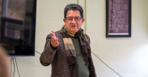 A man with dark hair wearing glasses, a gray sport coat and a brown scarf gestures with his right hand while speaking. (Photo by Keith Nordstrom)