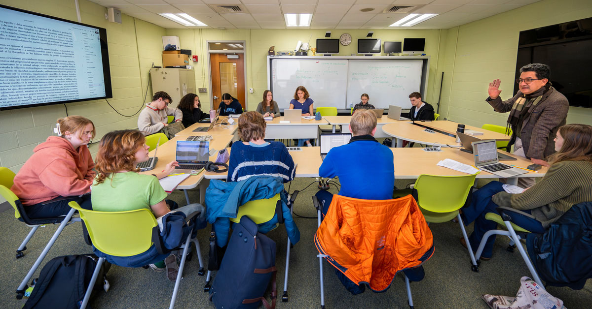 A male professor with dark hair wearing glasses and a dark sportcoat addresses a classroom of a dozen college students sitting at desks facing each other.