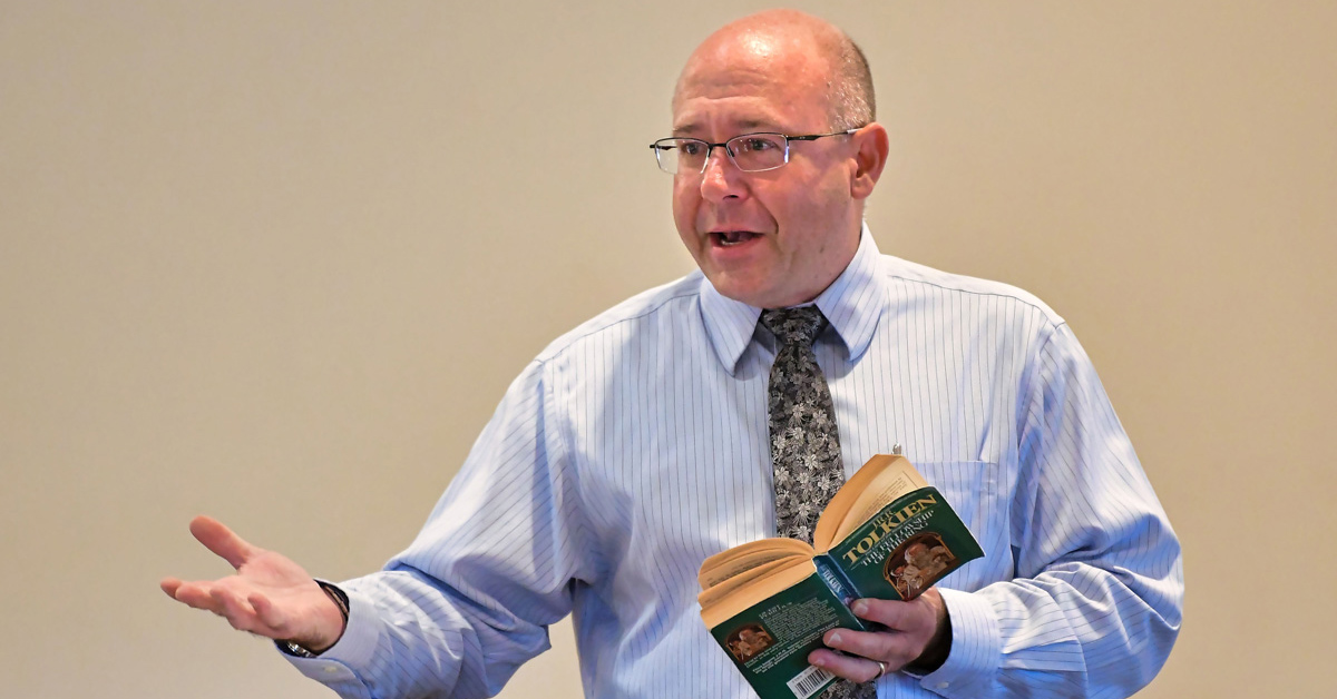 A balding white man wearing glasses and a light blue collared shirt with dark patterned necktie holds a Tolkien book in his left hand while gesturing with his right arm.