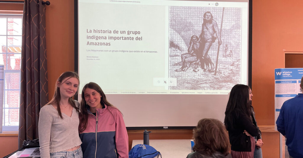 Two smiling young adult females stand in front of a projector screen with a black and white drawing of a pair of indigenous male hunters with one holding a staff. Text on the screen reads La historia de un grupo indigena importante del Amazonas
