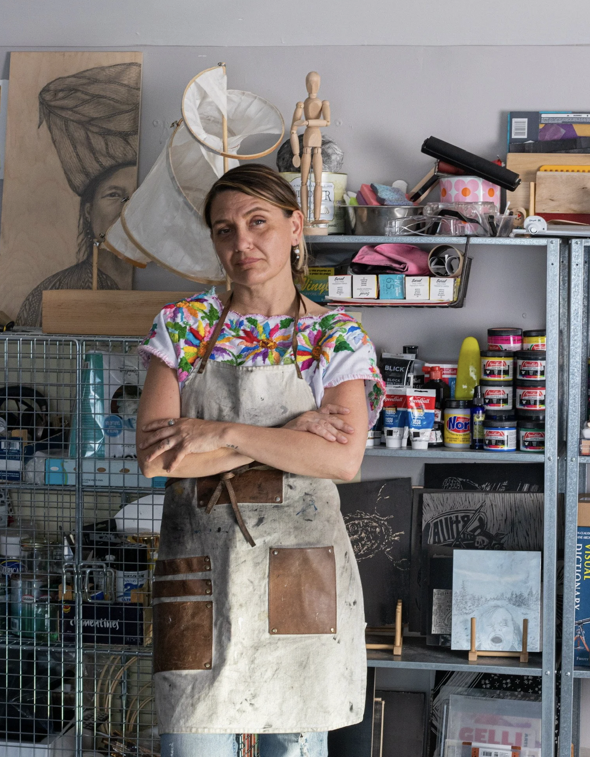 Artist Magda Leon standing in front of shelves of art supplies. Her arms are crossed and she is wearing a printmaking apron.