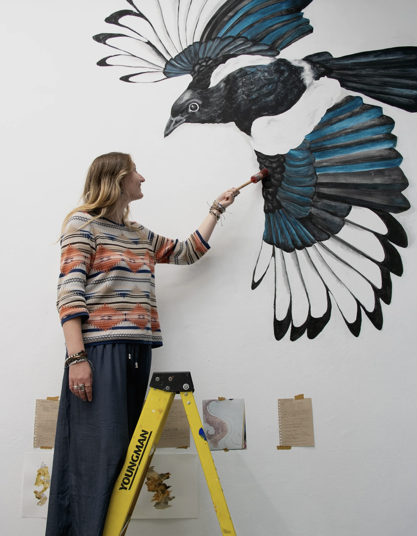 Artist Maia Hay standing on a yellow ladder with a paint brush in her left hand. Behind her is a large wall mural of a black, white, and blue bird with its wings outstretched.