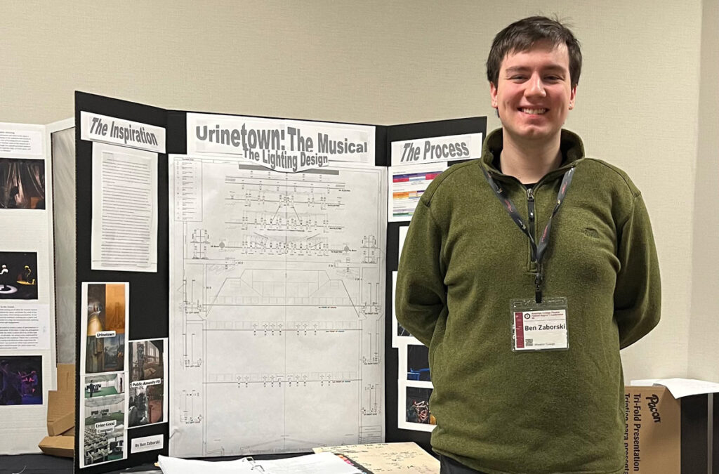 A smiling young man with dark hair wearing an olive green sweater stands in front of a posterboard display. The title reads Urinetown: The Musical!