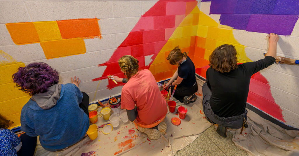Five individuals face away from the camera, sitting or kneeling, while painting the concrete walls of a staircase pink, orange and yellow.
