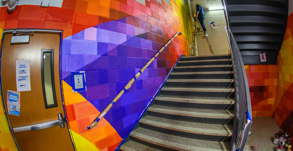 A variety of yellow, orange, red, blue and purple colors the walls of an internal staircase. An individual standing on a step ladder applies paint to a wall at the top of the stairs.