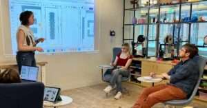Student stands before a slide displayed on a screen presenting an idea to an audience of fellow students and a professor.