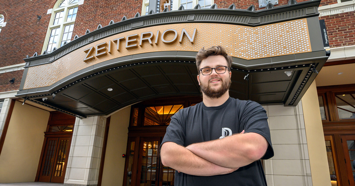A smiling young man with brown hair, a beard and a mustache wearing glasses and a black T-shirt folds his arms while standing in front of a building, with a sign reading Zeiterion above the entrance.