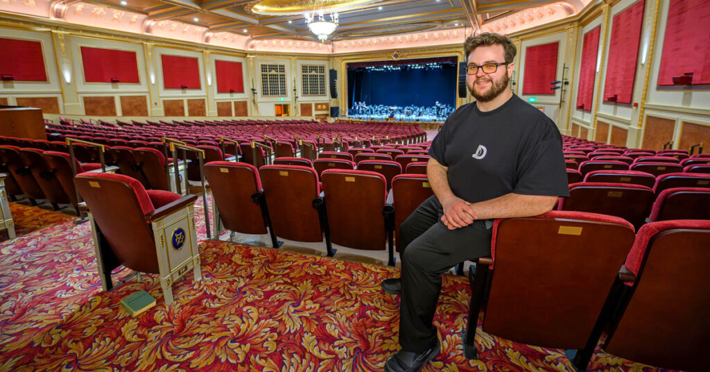 A smiling young man with brown hair, a beard and a mustache wearing glasses and a black T-shirt leans on a chair at the back of a theatre.