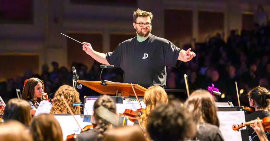 A smiling young man with brown hair, a beard and a mustache wearing glasses and a black T-shirt conducts an orchestra.