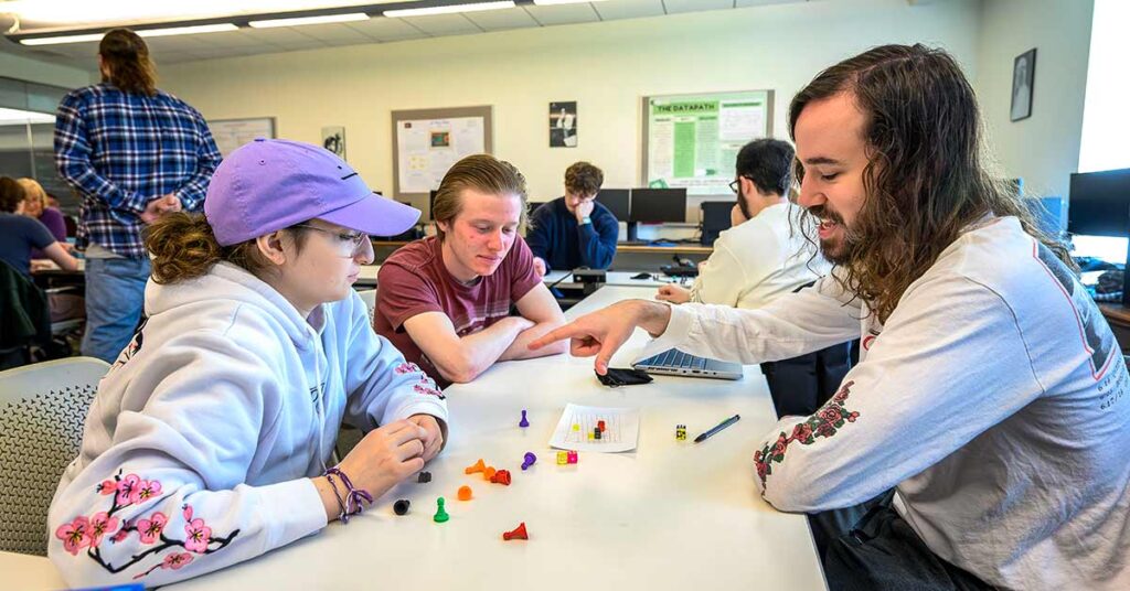 The image depicts a group of students engaged in a collaborative activity in a classroom setting. They are seated at a table with various colored game pieces in front of them. 