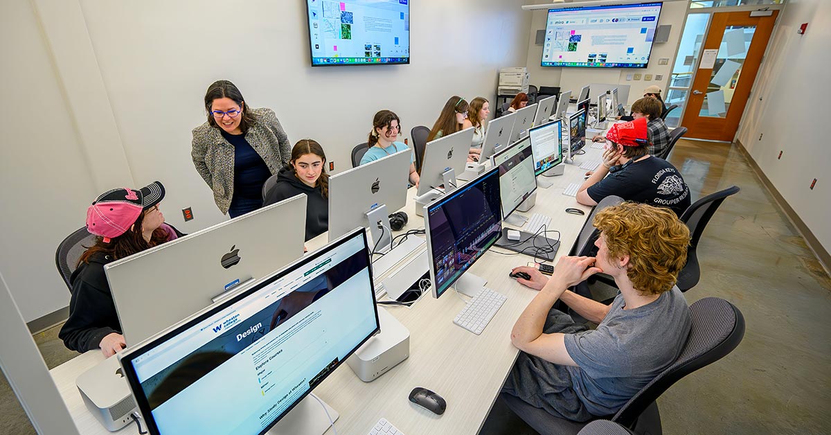 The image shows a classroom setting with several students seated at a long table, each working on desktop computers. The students are focused on their screens, which display various digital content. A teacher stands at the front of the room, engaging with a student. 
