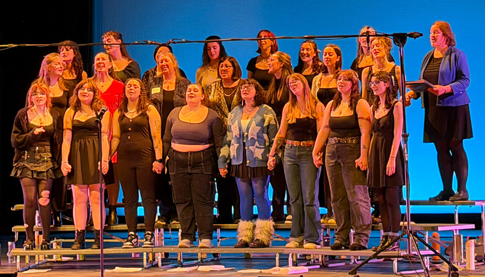 About 20 women sing together while standing on risers on a stage.
