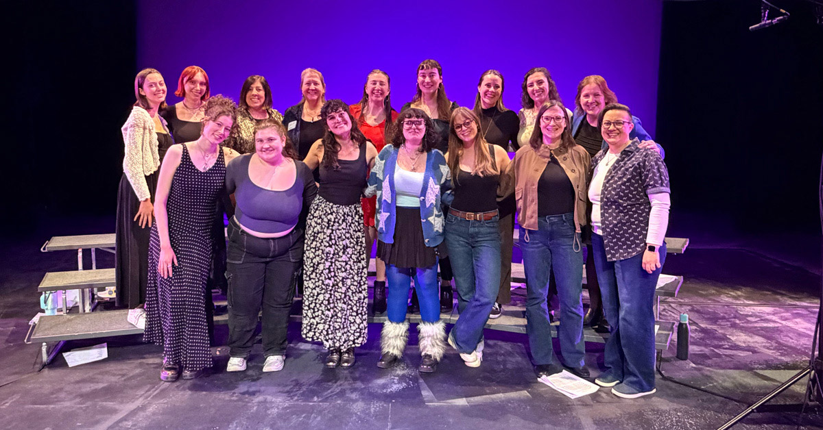 A group of 16 smiling women standing together on a stage.