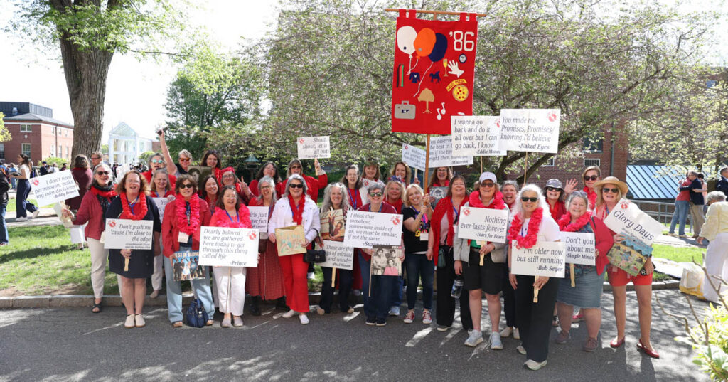 A large group of women, holding signs with lyrics from popular songs and a large decorated red flag, gather outside.