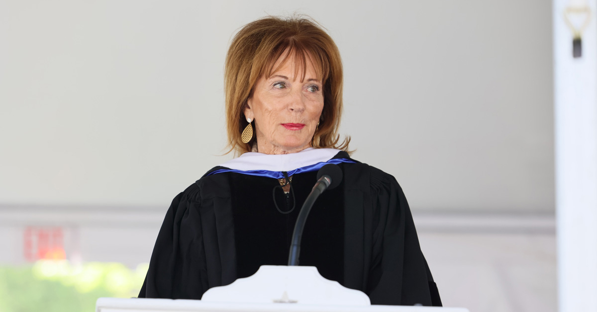 A woman with shoulder length brown hair wearing a black academic robe speaks from a podium.