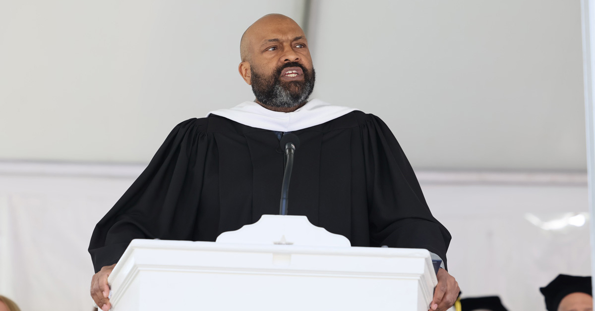 A bald man with a mustache and beard wearing a black academic robe speaks from a podium.