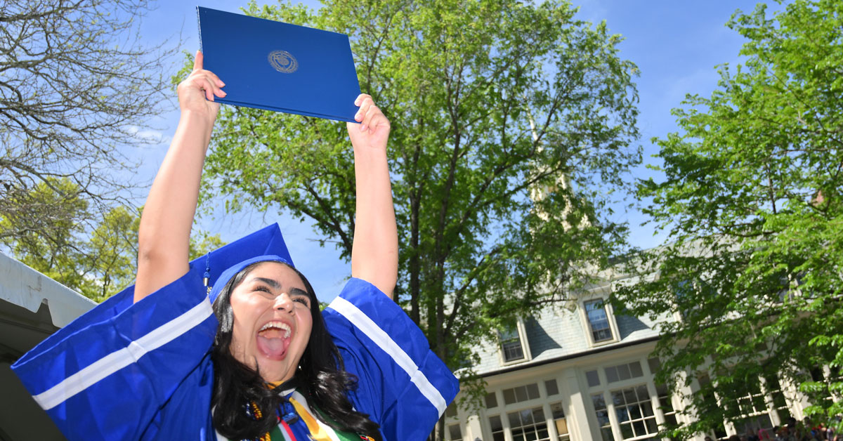 An excited woman wearing a blue academic robe and mortarboard proudly holds her college diploma over her head.