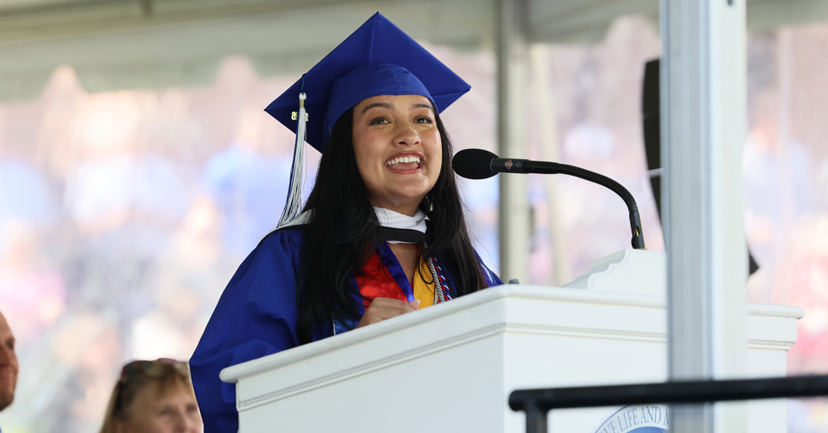 A young woman with long, straight black hair wearing a blue robe and mortarboard speaks from a podium.