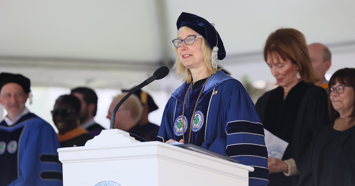 A woman wearing glasses, a black academic cap and a blue academic robe with black accents speaks from a podium.
