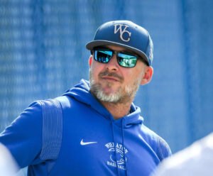 A man with a graying mustache and beard wearing a royal blue hooded sweatshirt, sunglasses and a baseball cap with WC on it.