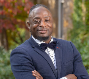 A man wearing a navy blue suit and bowtie, folding his arms.