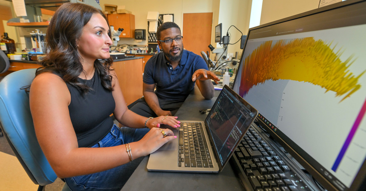 The image depicts a collaborative scene within a laboratory setting. Two individuals, a woman and a man, are intently focused on a computer screen displaying a complex, color-coded 3D visualization. The scene suggests a scientific or technical discussion.