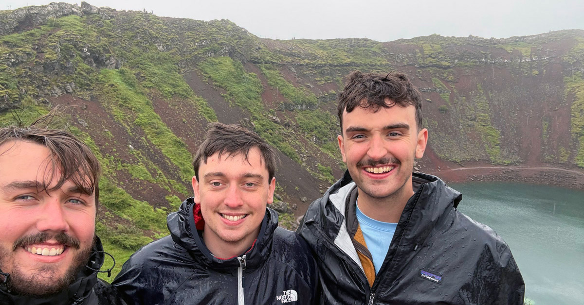 Three men pose for a selfie in rainy weather beside a turquoise volcanic lake surrounded by steep, moss-covered rocky terrain. All three are wearing waterproof jackets and smiling at the camera.