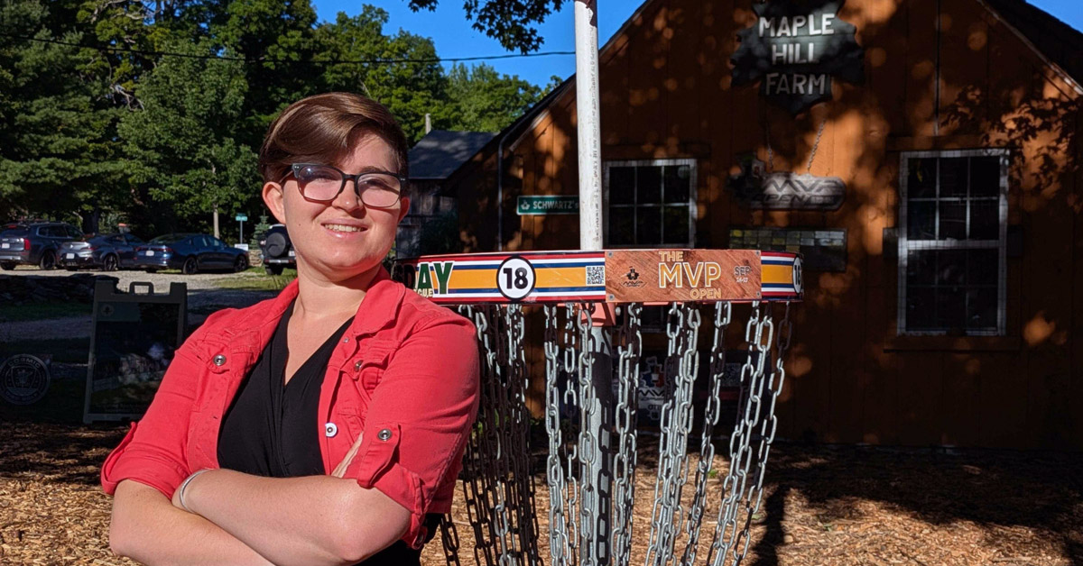 A smiling individual wearing glasses and a red shirt over a black top crosses their arms while leaning against a disc golf basket on a sunny day. A Maple Hill Farm sign can be seen on a red building among shady trees in the background.