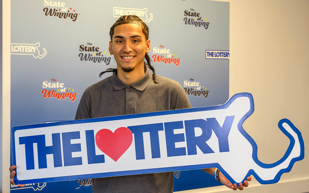 The image depicts a young man smiling and holding a large, blue, cardboard cutout of “The Lottery” logo. The logo features a heart within the outline of the state of Massachusetts. Behind him is a wall adorned with multiple “State of Winning” lottery posters.