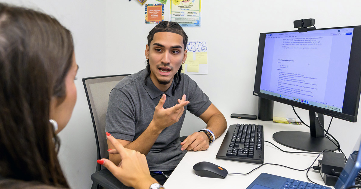 The image depicts a professional meeting in an office setting. A young man with dark hair is seated in a gray office chair, gesturing emphatically while speaking to a woman who is seated beside him. Both individuals are positioned at a white desk that is equipped with a computer monitor, a keyboard, and a mouse. A computer laptop is also visible on the desk.