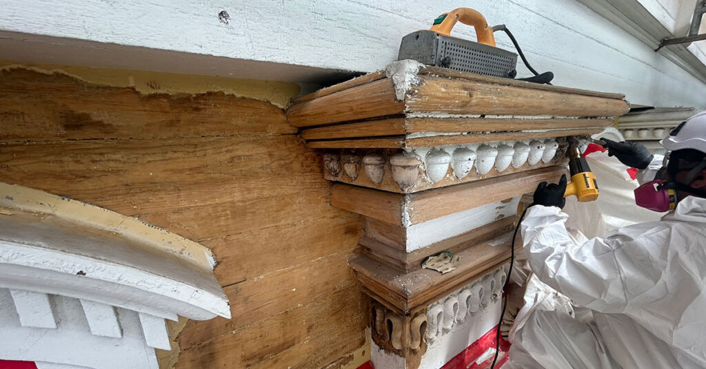 A worker wearing protective clothing and a respirator uses an infrared tool to remove paint from Mary Lyon Hall’s decorative woodwork.